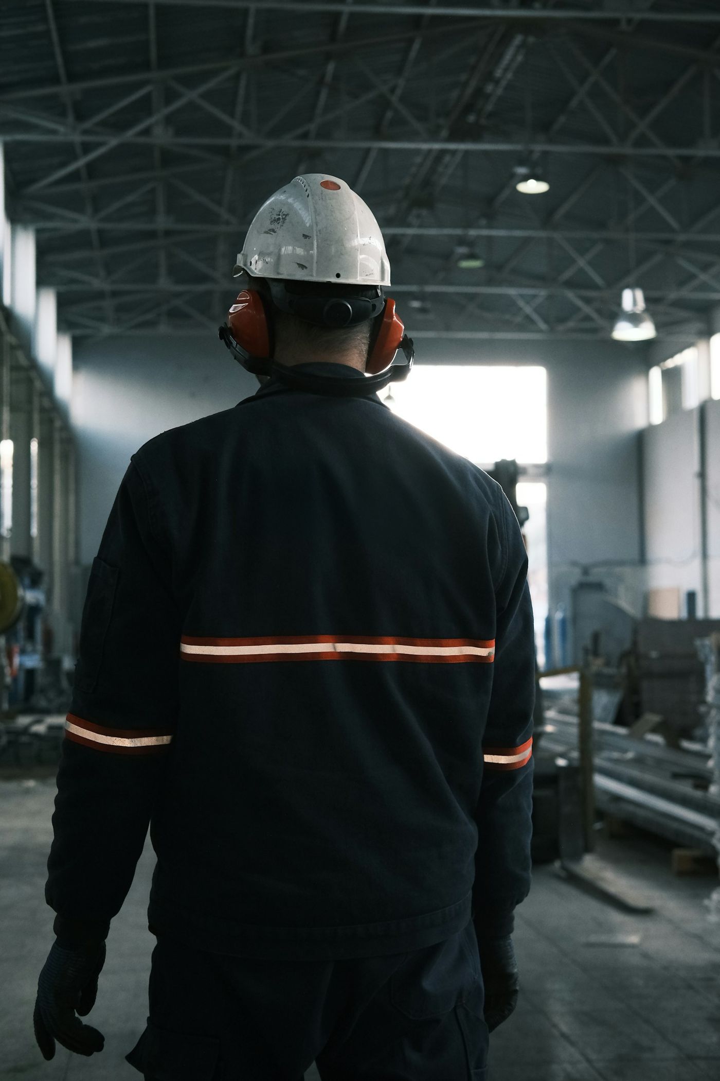 Worker in a white hard hat, red earmuffs and dark workwear with reflective tape, walking through a factory