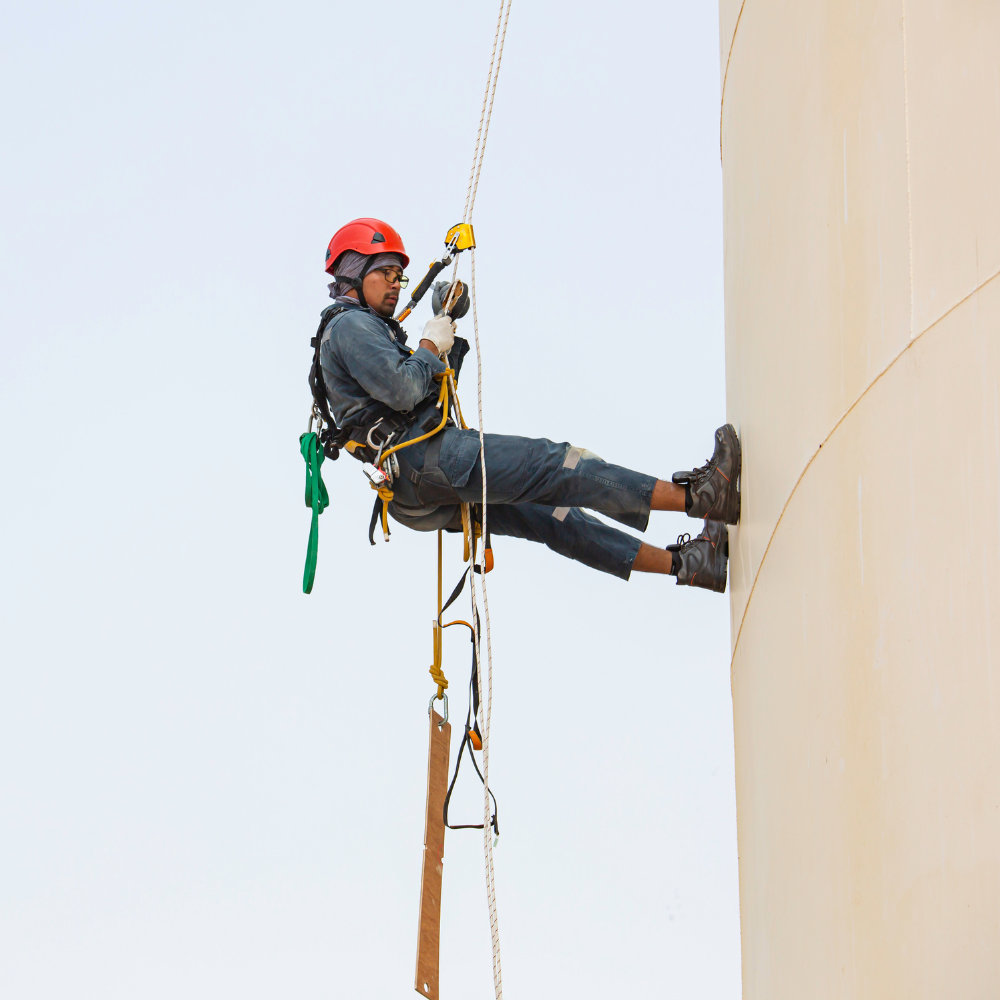 Rope-access worker in a red helmet and full-body harness working on the outside wall of a tall tank