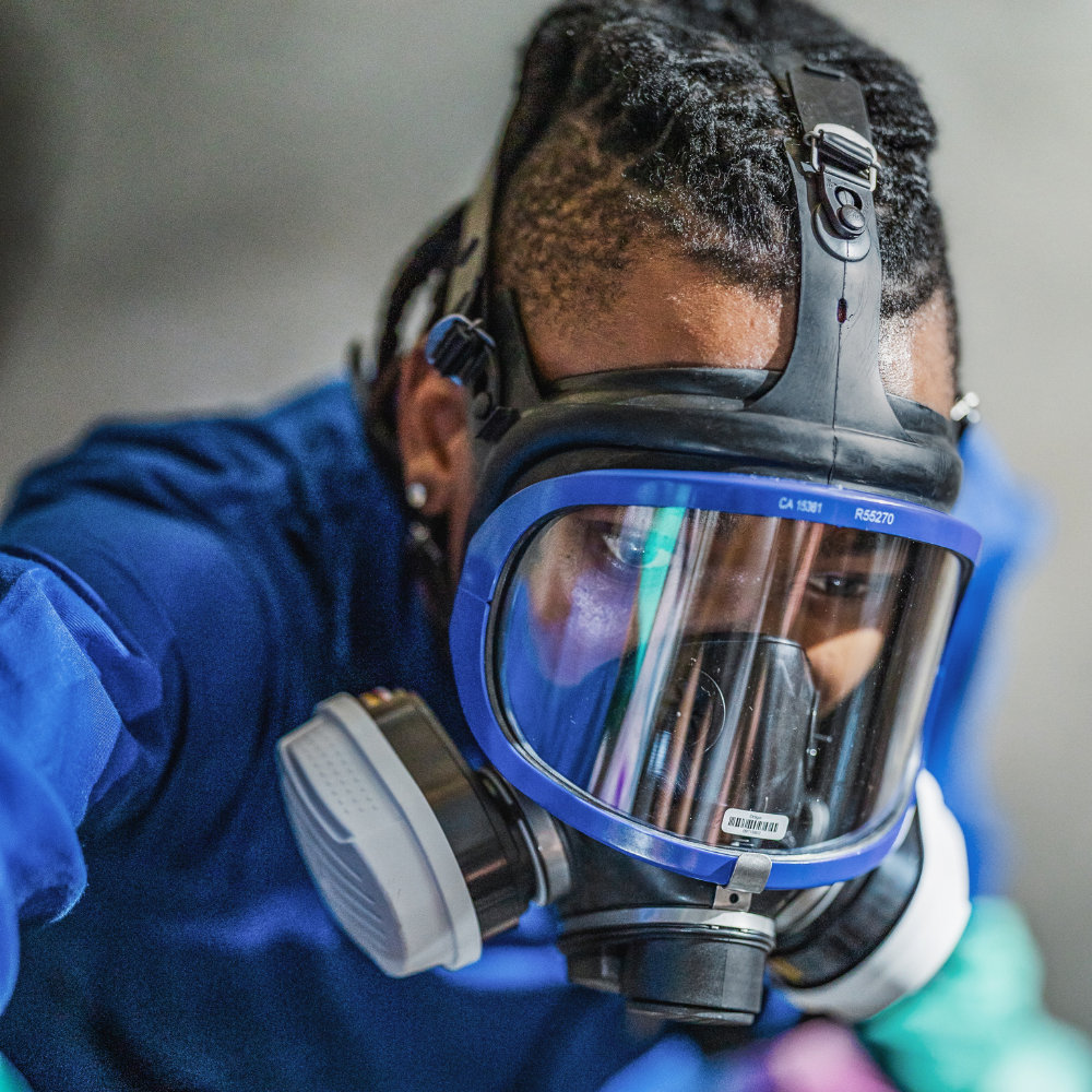 Worker in a blue coverall wearing a full-face respirator mask with a clear visor and twin filters
