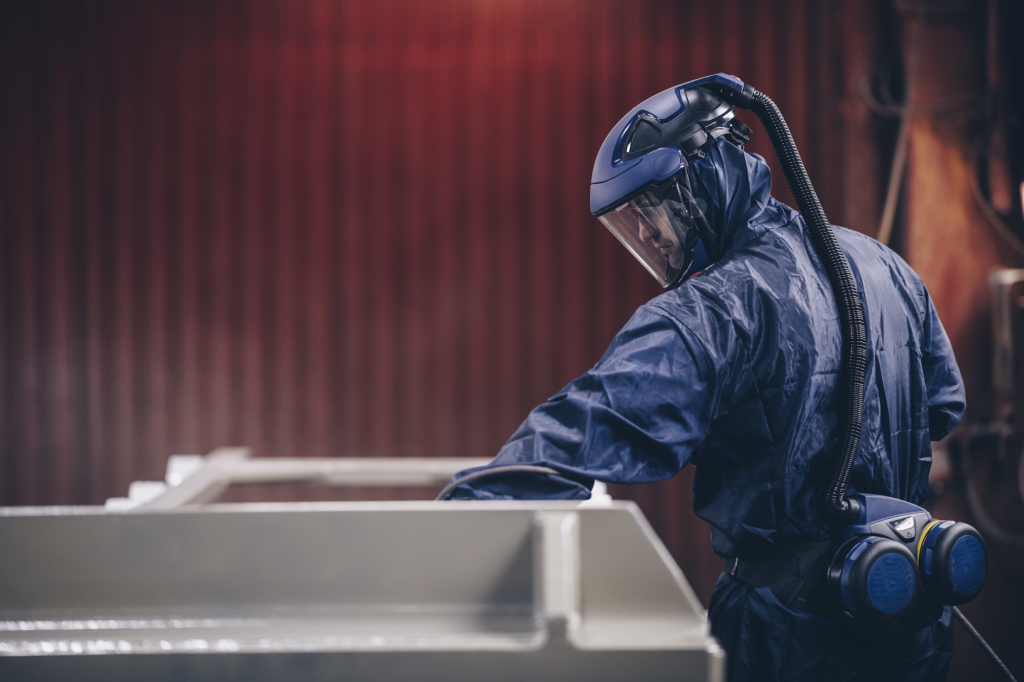 sundstrom-powered-air-respirator-hero Worker in a blue disposable coverall wearing a Sundstrom powered air respirator: helmet with flip-up visor and breathing hose to a belt-mounted fan unit with twin filters
